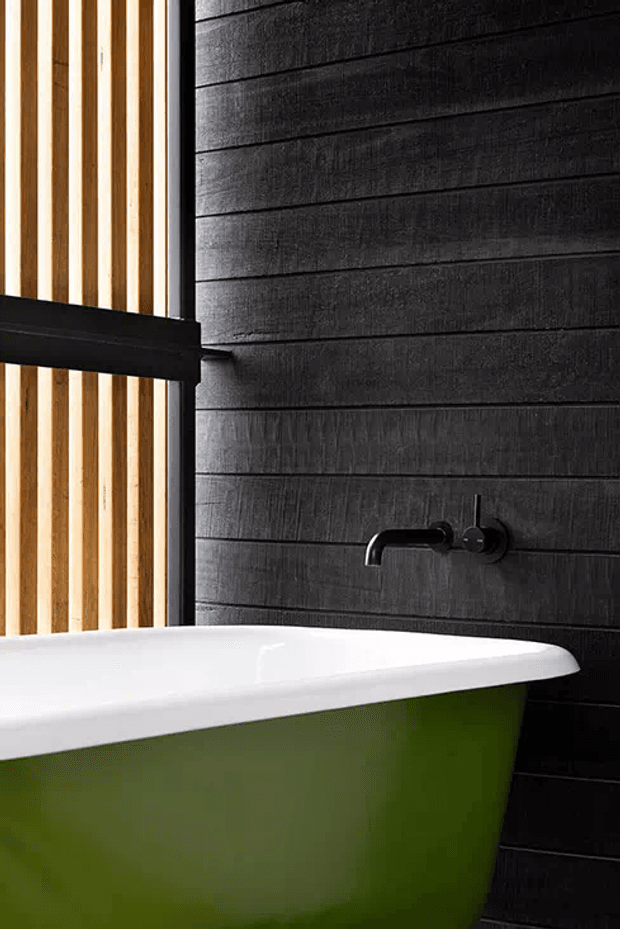 Close-up of a contemporary bathroom featuring a freestanding white-rimmed bathtub with a green exterior, matte black wall-mounted tapware, dark horizontal timber wall panelling, and a vertical light timber slatted screen beside the bath.