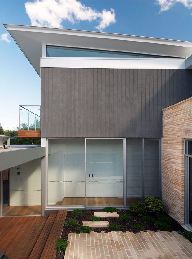 Exterior view of a contemporary multi-level residence centred on a sharply projecting roof, dark vertical cladding, extensive glazing, and a contrasting light brick wall. A timber deck, glazed balcony, and landscaped courtyard with brick paving occupy the foreground.