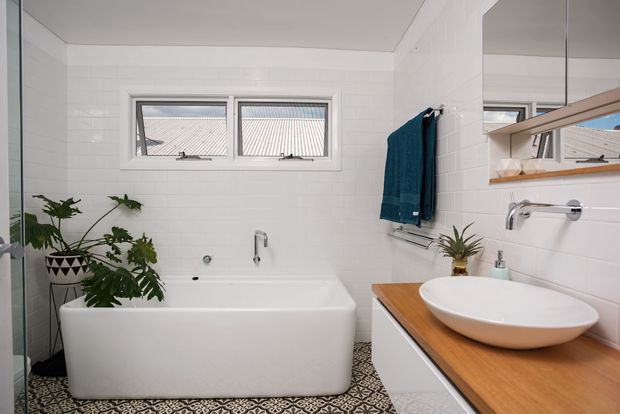 Bright contemporary bathroom with a freestanding white bathtub, white rectangular wall tiles, patterned floor tiles, and a timber vanity benchtop with a white vessel basin. A large mirrored cabinet, chrome tapware, narrow horizontal windows, towel rails, and indoor plants complete the space.