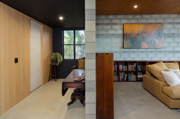 Interior view of a contemporary living room and adjoining hallway or study zone, divided by a prominent exposed concrete-block structural wall. The living area has a grey polished concrete floor, built-in dark timber bookcase, warm timber ceiling, large abstract artwork, and a tan sectional sofa. The adjoining space features extensive light timber wall panelling, a concealed flush door, dark ceiling, desk, plant, and black-framed window.