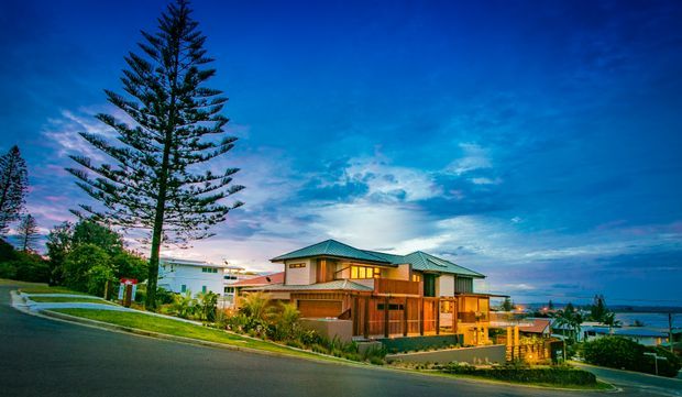 Twilight exterior view of a multi-level contemporary coastal residence on a sloping suburban site. The home features a low-pitched hipped roof, extensive warm-lit glazing, timber-toned cladding and balconies, with a large Norfolk Island pine and distant water visible in the background.