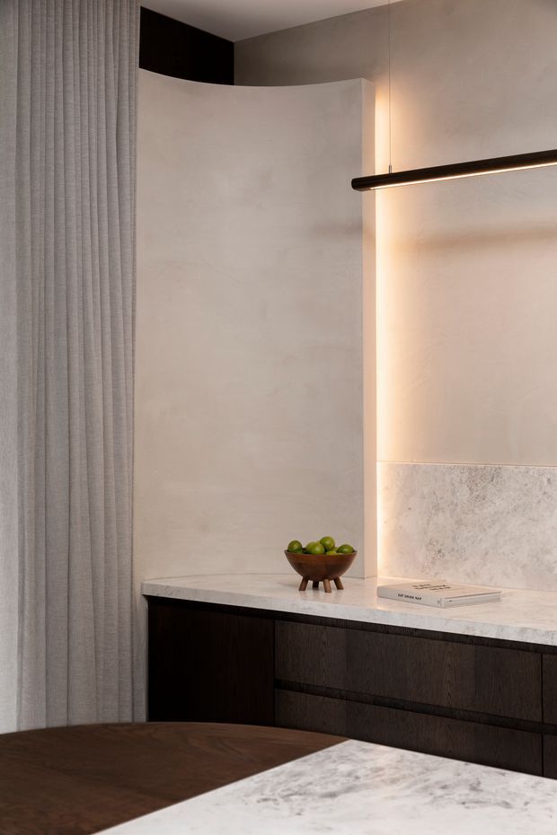 A refined contemporary kitchen interior featuring dark timber cabinetry, a pale stone benchtop and splashback, softly textured plaster walls, full-height grey drapery, and a slender black linear pendant light. A small timber fruit bowl and books sit on the rear counter, while the foreground shows a dark timber dining surface beside a marble-like island or benchtop. Warm concealed lighting creates a calm, luxurious atmosphere.