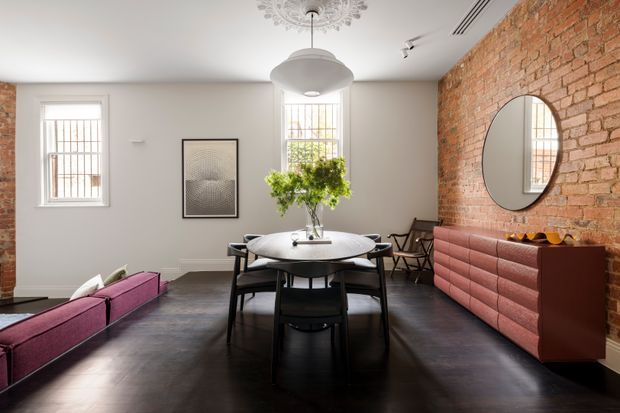 A bright, loft-like dining and living space with a round dining table and black chairs centered beneath a pendant light. The room features exposed red brick walls, dark timber flooring, white painted walls, tall sash-style windows, and a distinctive pink-red upholstered sideboard along the brick wall. A magenta sofa is partially visible in the foreground, with a round wall mirror, framed artwork, indoor greenery, and exposed brick adding character.
