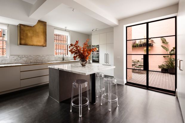 Contemporary kitchen with a large marble-topped island, dark integrated cabinetry, a bronze-toned rangehood feature, stainless appliances, and transparent bar stools. Black-framed glazed doors open to a small brick-paved courtyard with planted greenery. The room has a bright, refined palette with strong black-and-white contrast and warm metallic accents.