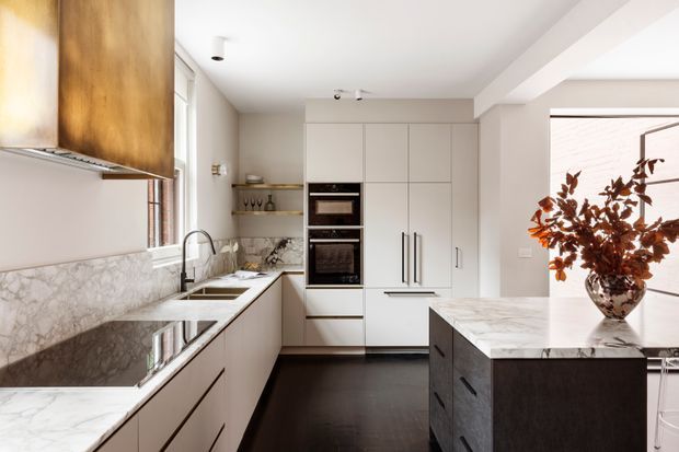 Bright contemporary kitchen with white full-height cabinetry, integrated double ovens, marble-look splashback and benchtops, a dark island with waterfall edge, dark timber flooring, and a sculptural timber rangehood. A large window and decorative vase add warmth to the refined space.