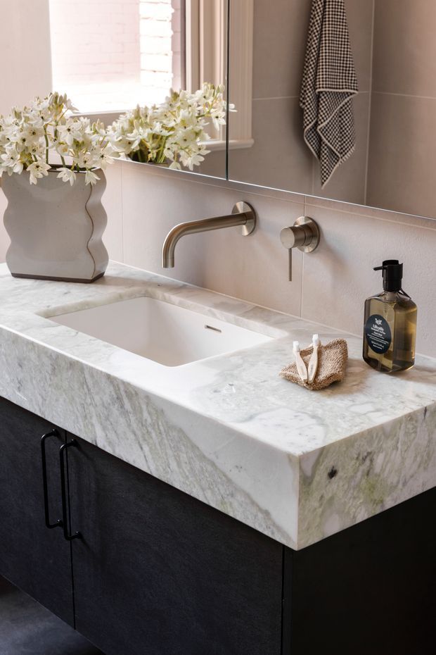 Close-up of a refined bathroom vanity featuring a thick white-veined natural stone benchtop with an integrated rectangular basin, wall-mounted brushed metal tapware, dark matte cabinetry with black pulls, a large mirror, warm taupe wall tiles, and a decorative vase of white flowers.