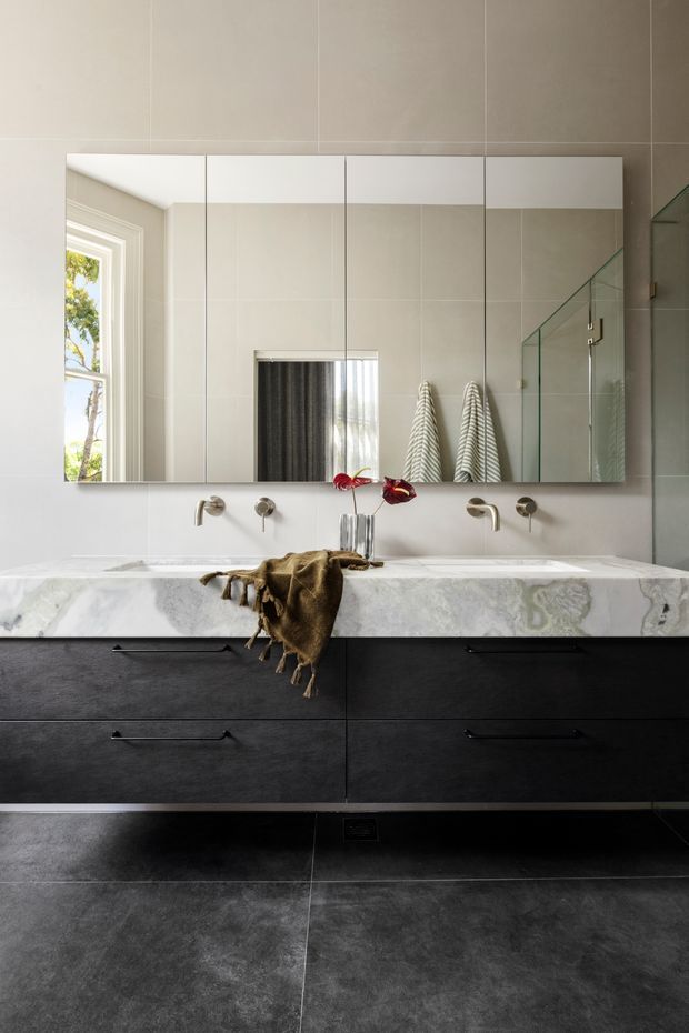 A refined contemporary bathroom vanity with a wide double basin, white marble benchtop, dark textured drawer cabinetry, wall-mounted tapware, large mirrored cabinet, and a frameless glass shower enclosure. Large-format grey wall and floor tiles create a restrained, monochromatic scheme, with natural light entering through a side window.