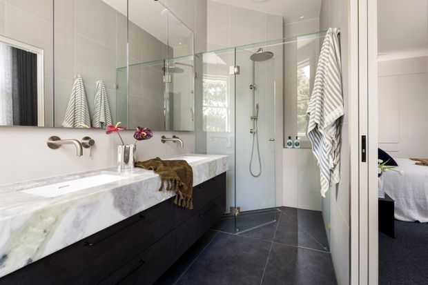 Bright contemporary bathroom with a long double vanity featuring a dramatic white-and-grey marble benchtop and dark integrated cabinetry. A frameless glass walk-in shower occupies the centre, with large mirrored wall panels, pale tiled walls, dark large-format floor tiles, and brushed metal wall-mounted tapware. A bedroom is visible through the open doorway at right.