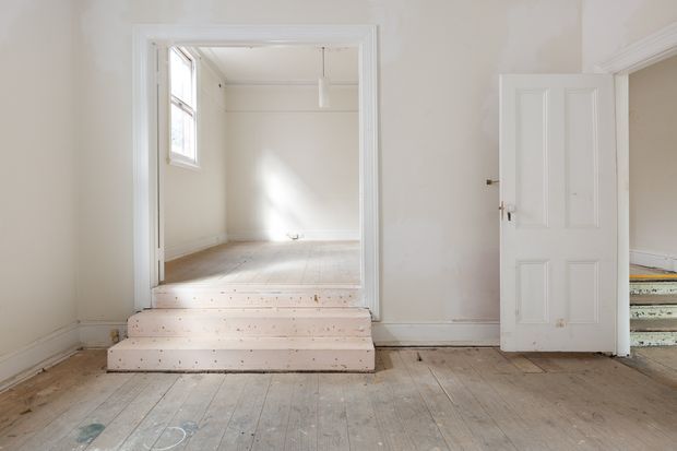 Bright, largely empty traditional interior with white-painted walls, timber floorboards, decorative ceiling cornices, and a raised adjoining room accessed by several broad steps. A white paneled door opens toward another room, creating a simple, worn architectural shell with abundant natural light.