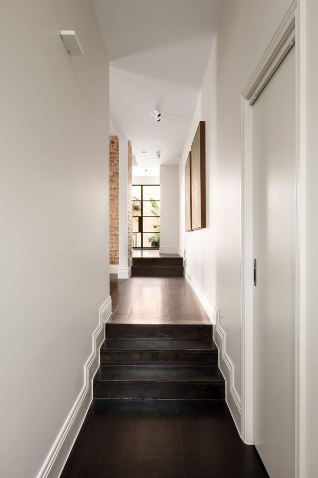 Bright narrow interior hallway with a short flight of dark timber stairs, white painted walls and decorative skirting and trim. Exposed brickwork is visible along the left side, while framed artwork and a white door line the right wall. The corridor continues toward black-framed glazing and indoor greenery at the far end.