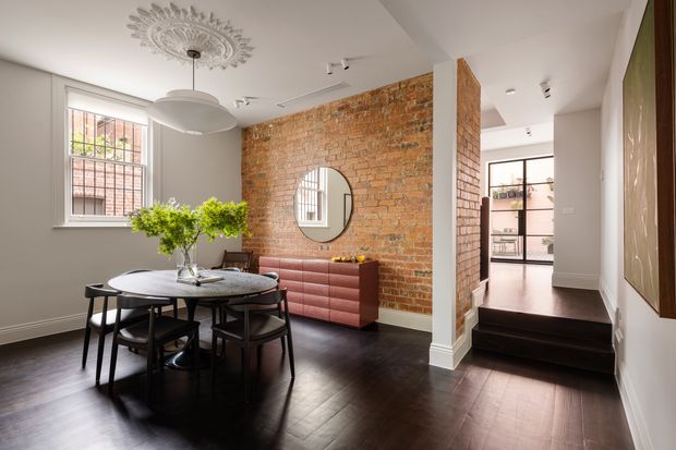 A refined dining area with a round dark timber dining table and black chairs, set against a white-painted interior and exposed red brick feature wall. A blush-pink ribbed sideboard sits below a circular mirror, while a sculptural pendant and ornate ceiling medallion add decorative character. Dark timber flooring leads to a raised hallway or adjoining space with black-framed glazing and built-in shelving.