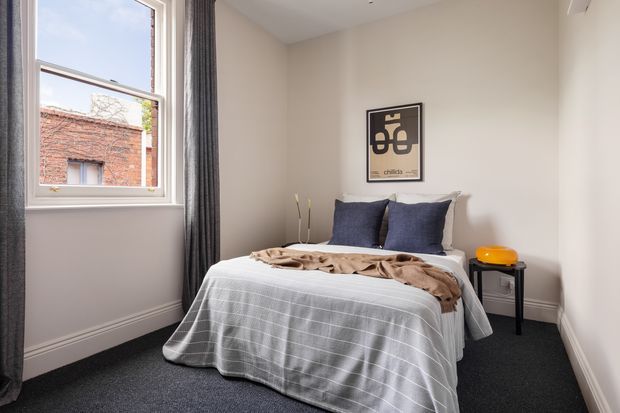 Compact, simply styled bedroom with a double bed dressed in a white textured cover and dark blue cushions, positioned beside a large window with dark grey curtains. The room has cream-painted walls, white skirting boards, dark carpet, a small black side table with an orange lamp, and a framed graphic print above the bed.