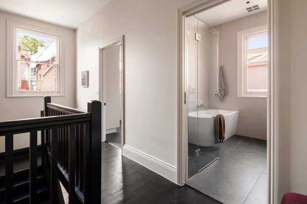 A bright upstairs hallway and landing with dark timber flooring and black stair balustrade, looking toward an open bathroom. The bathroom has large grey floor tiles, pale wall tiles, a freestanding white bathtub, clear glass shower enclosure, and a window. White painted walls, decorative skirting boards, and traditional window and door trim are visible throughout.