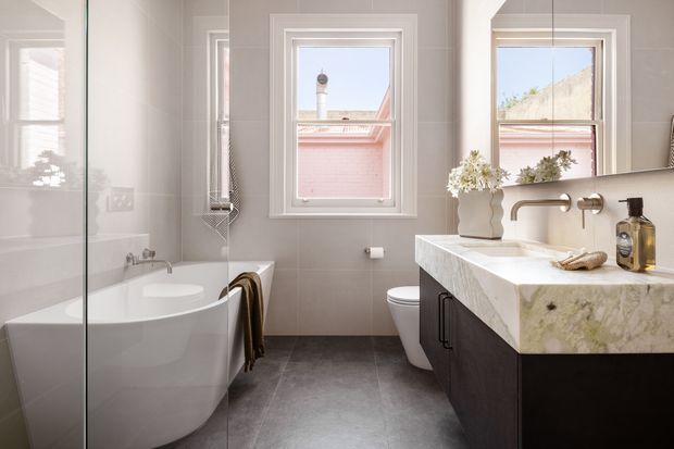 Bright, refined bathroom with a freestanding white bathtub beside a frameless glass shower screen, a wall-mounted toilet, and a dark timber vanity topped with a thick pale stone benchtop. Neutral tiled walls, grey flooring, white-trimmed windows, brushed metal tapware, and soft botanical styling create a clean contemporary look.