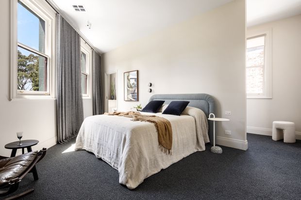 Bright, refined bedroom with a large upholstered bed, layered neutral bedding, dark blue cushions, and a soft brown throw. Tall windows with grey full-length curtains provide abundant natural light, while dark carpet, white walls, minimalist artwork, and restrained furnishings create a calm contemporary interior. A small adjacent sitting area is visible to the right.