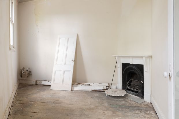 An empty, weathered interior room undergoing renovation. The space has aged timber floorboards, plain off-white plaster walls, a tall narrow window, a detached white paneled door leaning against the wall, exposed skirting and plumbing near the floor, and a traditional black cast-iron fireplace with a simple white mantel.