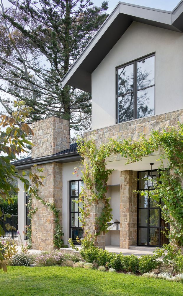 Exterior view of a multi-level contemporary home with a sharply pitched gable roof, white rendered walls, extensive pale natural-stone cladding, and dark-framed glazing. A covered entry or outdoor terrace is framed by stone columns and climbing greenery, with landscaped garden beds and lawn in the foreground. Tall mature trees surround the house.