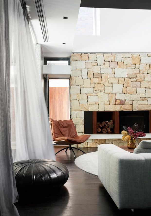 A refined contemporary living room with a large natural irregular-stone fireplace wall, dark timber flooring, floor-to-ceiling sheer curtains, and a built-in wood storage niche. The room includes a sculptural brown leather lounge chair, black leather ottoman, pale upholstered sectional sofa, and a round light rug. High ceilings, recessed lighting, and a tall glazed opening create an airy yet warm atmosphere.