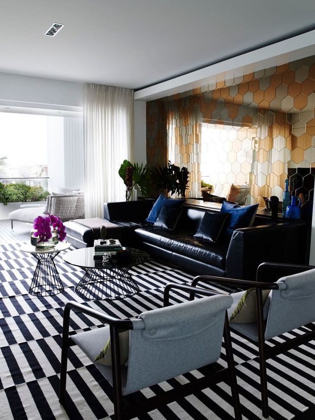 A bright, sophisticated living room with a black leather sofa, pale upholstered lounge chairs, geometric wire-frame coffee and side tables, and a bold black-and-white striped rug. The focal wall features a reflective honeycomb-patterned metallic tile or panel installation in warm bronze, gold, and grey tones, with sheer curtains and abundant natural light.