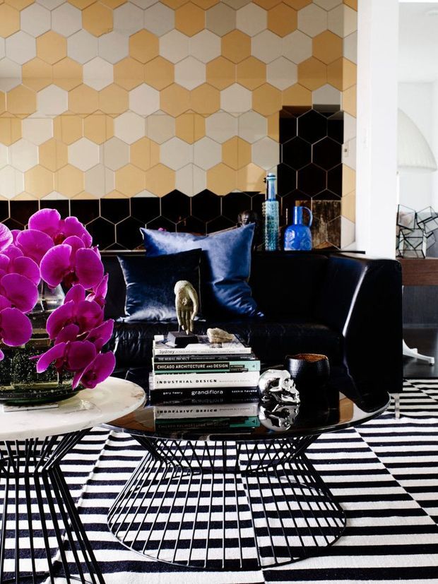 A bold, eclectic living room featuring a black leather sofa, deep blue cushions, a black wire-frame coffee table, and a striking black-and-white geometric rug. The wall behind the sofa is covered in a honeycomb-patterned tile or panel finish in white, beige, mustard, and black. Purple orchids, blue glass accessories, books, and sculptural objects add vivid accents. A partial dining or workspace area is visible through the opening on the right.