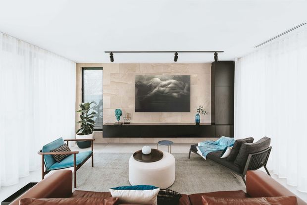 Bright contemporary living room with full-height sheer curtains, a textured pale stone feature wall, and a sleek black built-in media console. The room includes a brown leather sofa in the foreground, black-framed lounge seating with turquoise cushions, a rounded white ottoman, a light rug, indoor plants, and a linear ceiling track light.