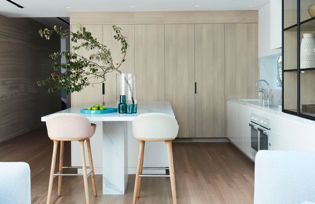 A bright, contemporary open-plan kitchen with a pale timber full-height cabinetry wall, white island and benchtops, integrated appliances, and timber-legged bar stools. A dark timber-toned feature wall contrasts with the light cabinetry and warm timber flooring. Black-framed glazed shelving and a leafy branch add visual interest.