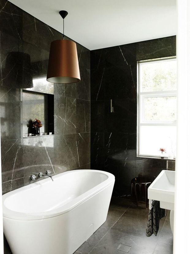 A refined bathroom featuring a freestanding white bathtub, dark charcoal marble-look wall and floor tiles with prominent white veining, a white basin vanity, a large window, and a striking copper pendant light. A mirrored recessed wall area adds depth to the compact space.