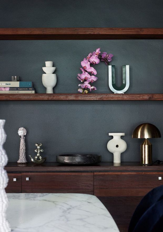 A close-up interior vignette featuring dark timber floating shelves and a matching low cabinet against a deep charcoal-blue textured wall. The shelves display books, sculptural ceramics, a pink orchid arrangement, a metallic U-shaped object, decorative figurines, a dark stone bowl, and a brass dome table lamp. The foreground includes the corner of a white marble-topped table or island. The styling is refined, moody, and contemporary with warm wood and metallic accents.