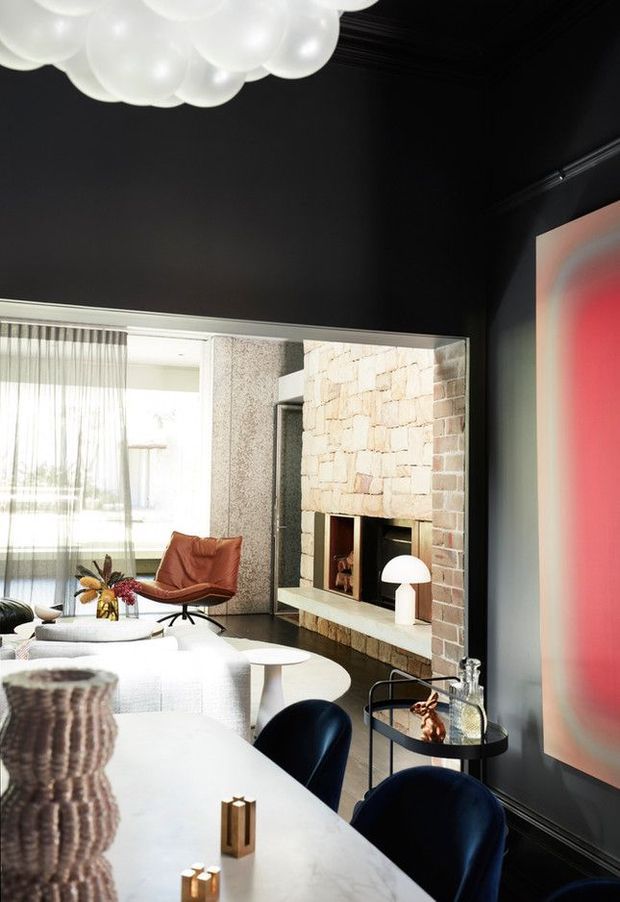 A sophisticated open-plan living and dining space with a dramatic black ceiling and walls, a large pale stone-clad fireplace, and expansive glazed doors with sheer curtains. The room features a contemporary white sofa, sculptural coffee tables, a warm leather lounge chair, dark blue dining chairs, a pale dining table, a sculptural white pendant light, and a vivid red artwork or feature panel. The overall design combines strong modern architectural elements with refined mid-century-inspired furnishings.