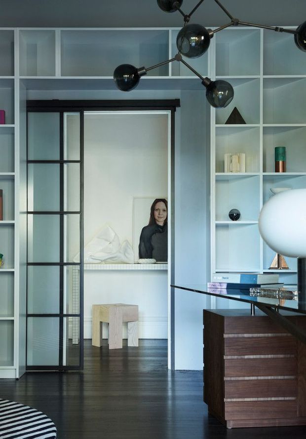 A refined interior view centered on a black-framed glazed sliding partition and an open bedroom beyond. Floor-to-ceiling white built-in shelving spans the wall, displaying books and objects, while dark timber flooring, a walnut-toned chest of drawers, and a sculptural black multi-arm pendant create a sophisticated mid-century-inspired scheme. A pale upholstered bed and small light timber stool are visible in the adjoining room.