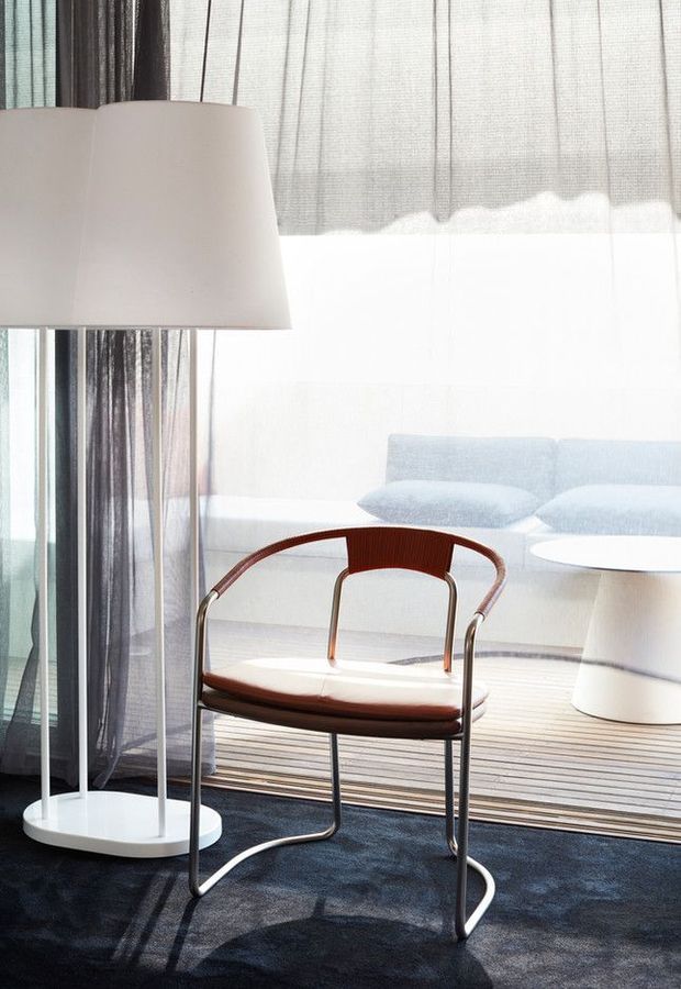 Bright contemporary living room with a sculptural white floor lamp, sheer layered curtains, pale blue-white sofa and rounded side table. A polished dark timber and metal lounge chair sits in the foreground on a dark blue rug, with strong natural light filtering through large glazing.