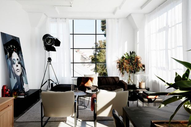 Bright, high-ceiling living room with expansive black-framed grid windows and sheer white curtains. The space contains a dark sofa, two cream lounge chairs, a coffee table, side tables, a large indoor plant, artwork, and studio-style lighting. Sunlight creates strong shadows across a neutral, contemporary interior.
