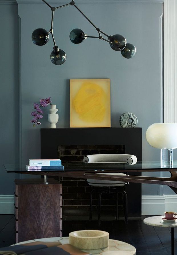 A refined living room or study vignette with dark blue-grey walls, detailed white ceiling and skirting mouldings, a black brick fireplace, and a sculptural dark timber desk spanning the foreground. A black branching globe pendant hangs overhead, while a modern desk lamp, books, artwork, ceramics, and a purple orchid add curated decorative details. The room has a sophisticated, moody contemporary character with strong mid-century-inspired furnishings.