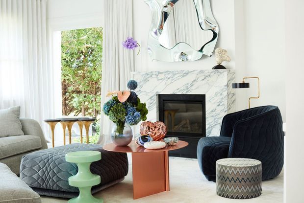 Bright, refined living room with a white marble fireplace surround, sculptural wall mirror, pale sheer curtains, a neutral sofa, dark blue upholstered armchair, quilted ottoman, and a sculptural coral-toned coffee table. Decorative floral arrangements and patterned occasional seating add colour and texture.