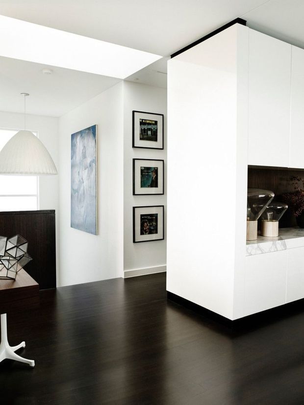 Bright contemporary interior showing a white kitchen volume with a marble benchtop and dark inset shelving, opening onto a dark timber-floored living or dining area. The space features crisp white walls and cabinetry, black trim accents, framed artwork, a sculptural pendant light, and minimalist furnishings.