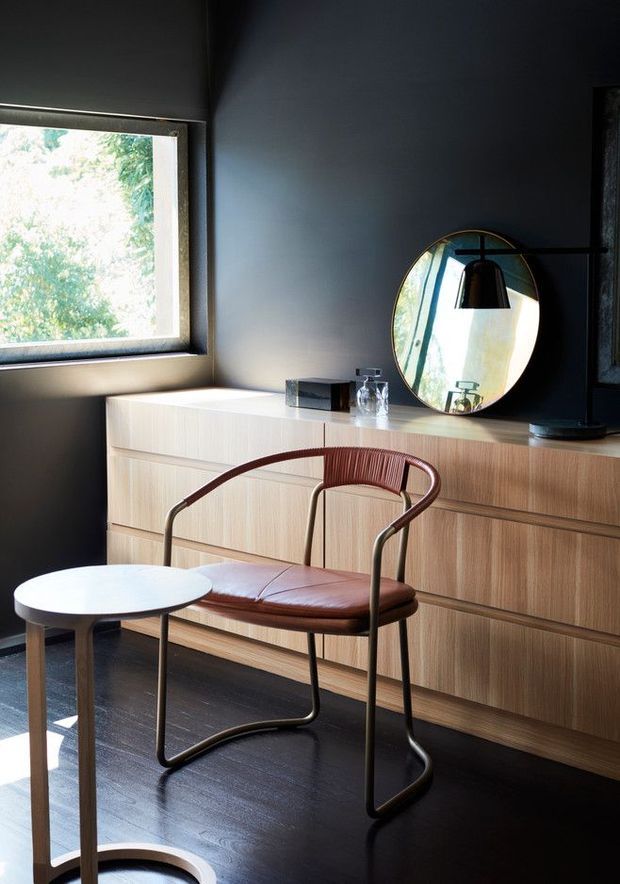 A dark, refined interior corner featuring a light timber built-in sideboard with horizontal drawers, a round wall mirror, and a sculptural leather-and-metal armchair beside a small round table. A large window brings in strong natural light, contrasting with the charcoal walls and dark timber floor.