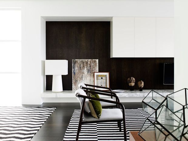 Contemporary living room with a dramatic dark timber feature wall and built-in cabinetry, light neutral walls and ceiling, and dark flooring. The room includes a white floor lamp, framed artwork, a low console, a cushioned dark timber armchair, striped rugs, and geometric glass-and-metal coffee tables. The overall composition is monochromatic with strong black-and-white contrast and warm wood accents.