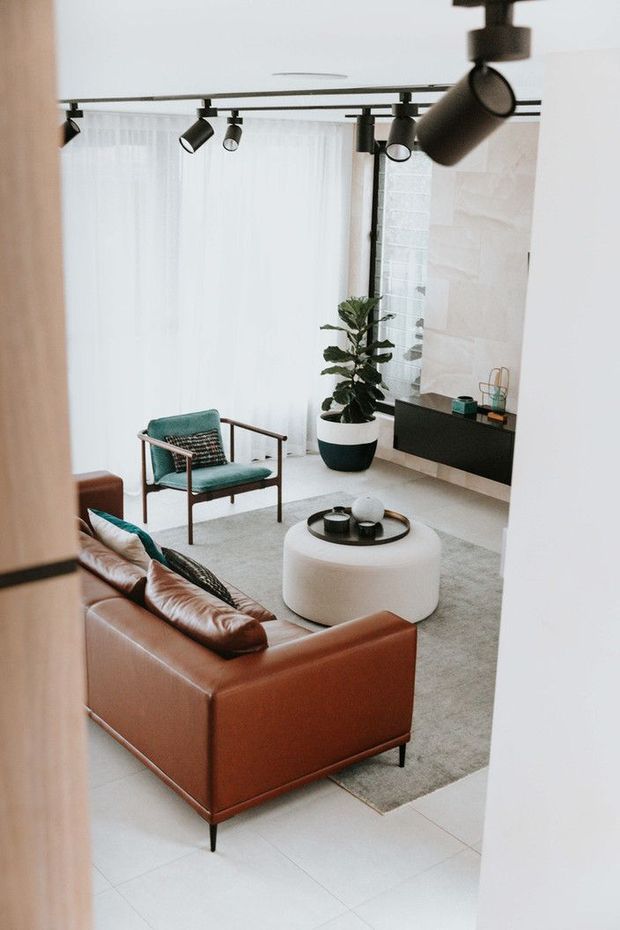 A bright, contemporary living room viewed from an adjacent doorway. The room features a cognac-brown leather sofa, teal upholstered armchair, rounded white coffee table, pale rug, large sheer-curtained window, black track lighting, a wall-mounted dark media console, indoor plant, and a light textured feature wall.