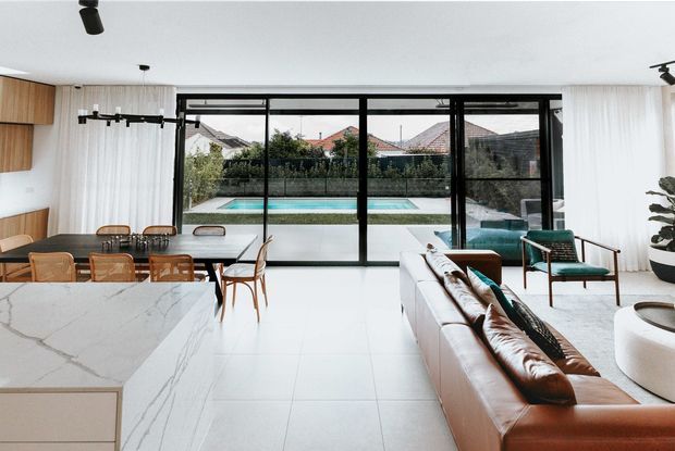Bright open-plan contemporary living and dining area with a large white stone kitchen island in the foreground, timber dining table and chairs to the left, and a leather sofa with green accent chairs to the right. Full-height black-framed glazing opens the room visually to a landscaped backyard and swimming pool. The space has white tiled flooring, white walls and curtains, timber cabinetry, track lighting, and a clean indoor-outdoor connection.