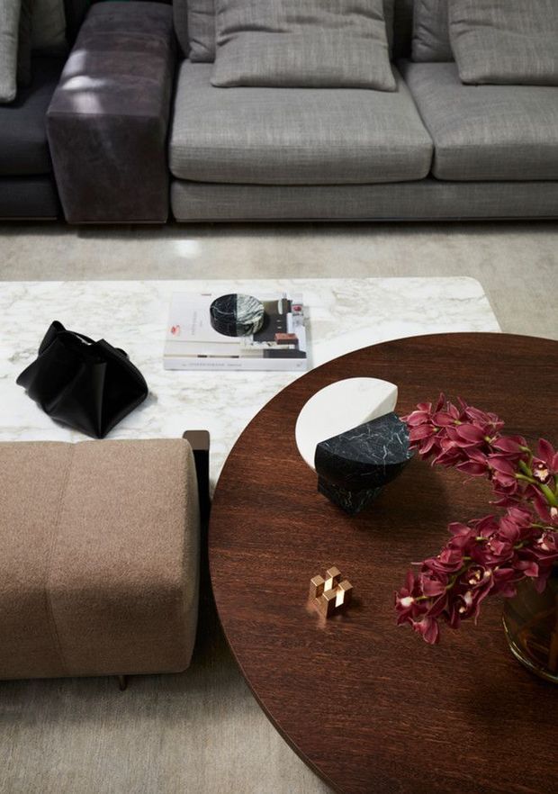 Close-up interior view of a refined living room seating arrangement. A dark grey sectional sofa sits behind a large round dark timber coffee table, accompanied by a tan upholstered ottoman and a white marble rectangular side table. The table includes sculptural black-and-white objects, books, a small gold decorative piece, and a floral arrangement. The palette is subdued and luxurious, with layered neutral upholstery and dark wood.