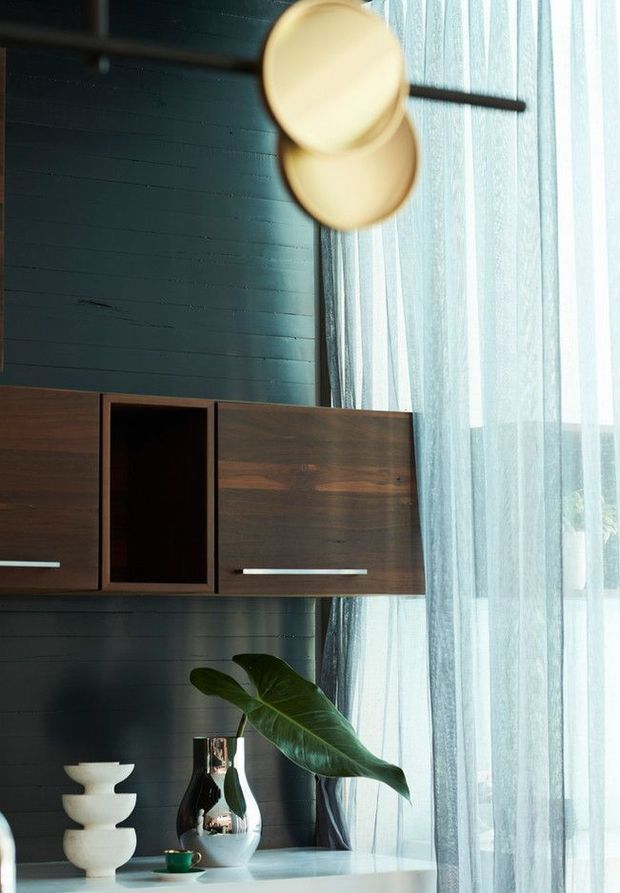A moody, refined interior vignette featuring a dark teal painted timber-panel wall, a wall-mounted dark timber storage cabinet with open shelving, and a pale countertop or console below. Sheer floor-to-ceiling curtains filter abundant daylight beside the cabinetry. A sculptural pendant light, white ceramic objects, reflective vase, and large green leaf provide decorative accents.