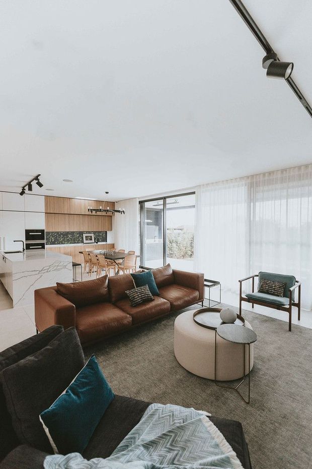 Bright open-plan contemporary living space with a warm modern aesthetic. The room combines a brown leather sofa, dark upholstered seating, teal cushions, a round upholstered coffee table, and a large neutral rug. Behind the living area is a minimalist kitchen with a white marble-look island, pale timber cabinetry, dark splashback, and integrated appliances. Floor-to-ceiling sheer curtains and large glazed doors bring in abundant natural light, while black track and pendant lighting add contrast.