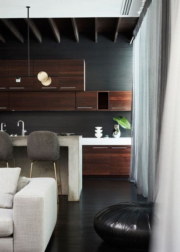 A refined open-plan kitchen and living area with dark timber cabinetry, a black timber feature wall, exposed ceiling rafters, and a pale concrete island with bar seating. Floor-to-ceiling sheer curtains line the glazed edge, while a light sofa and black leather ottoman create contrast against the dark flooring. Contemporary pendant lighting and restrained decor add a polished, moody feel.