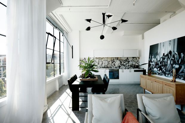 Bright open-plan apartment interior combining a dining area, compact white kitchen, and living space. The room features expansive black-framed windows with sheer curtains, a polished concrete-look floor, white cabinetry, a bold irregular stone mosaic splashback, a black sculptural pendant light, and a timber sideboard beneath a large monochrome artwork.