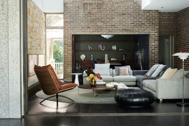 A sophisticated living room with exposed brick walls, a large built-in dark timber shelving and display unit, pale modular sofas, a sculptural leather lounge chair, a low coffee table, and a floor lamp. Natural light enters through tall glazing, contrasting with dark timber flooring and the warm, textured masonry.