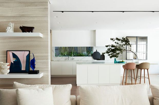 A bright, open-plan contemporary living area viewed from behind a white sofa, looking toward a minimalist kitchen with a large white island and two timber-and-upholstered bar stools. The room features pale timber wall panelling with integrated display shelves, white cabinetry, light flooring, indoor greenery, simple modern decor, and abundant natural light through large windows.