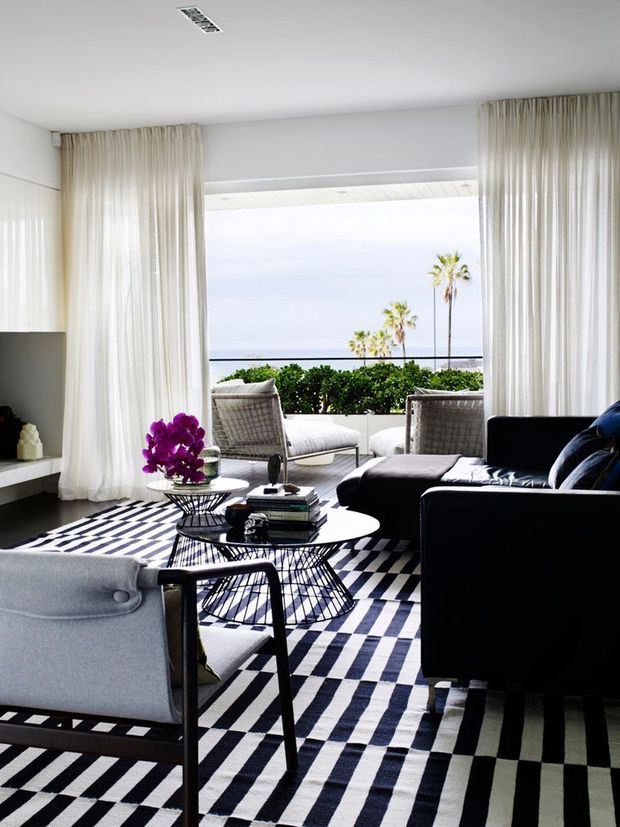 Bright contemporary living room opening onto a balcony or terrace with ocean views. The room features floor-to-ceiling sheer curtains, a black leather sectional sofa, light upholstered lounge chairs, sculptural black coffee tables, and a bold geometric black-and-white rug. Palm trees, balcony planting, and a dark balustrade are visible beyond the wide opening.