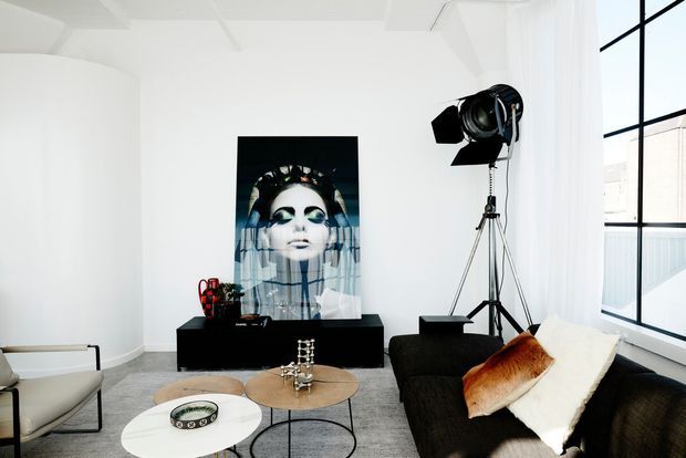 Bright, high-ceilinged contemporary living room with white walls, expansive black-framed glazing, a dark sectional sofa, layered coffee tables, sculptural lounge chairs, and a large fashion portrait artwork. A studio spotlight and tripod add an editorial, gallery-like character.