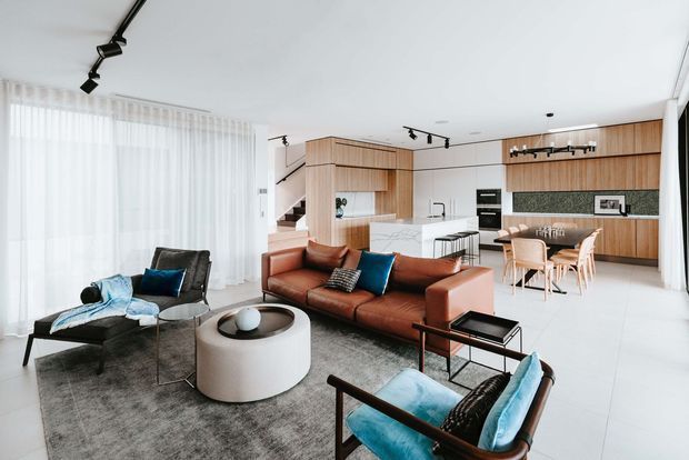 A bright, open-plan contemporary interior combining a living room, dining area, and kitchen. The living space features a cognac leather sofa, teal upholstered lounge chairs, a round sculptural coffee table, a large grey rug, and black track lighting. Behind it, a timber-lined kitchen with integrated cabinetry, a stone island, stainless fixtures, and a patterned backsplash opens to a light timber dining area.
