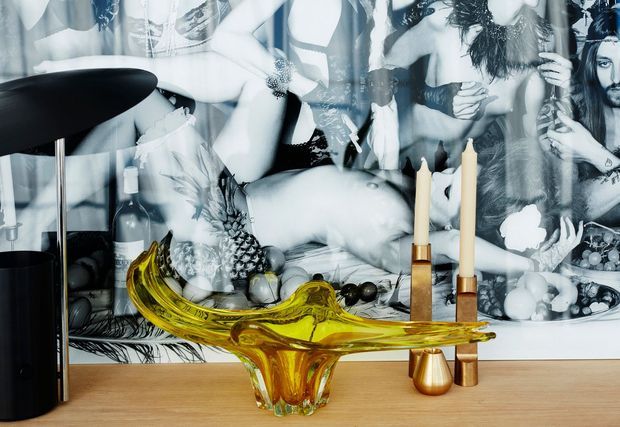 A moody, stylised interior vignette featuring a light timber console or sideboard topped with a sculptural translucent amber glass bowl, brass-toned candleholders with cream taper candles, fruit, and a dark table lamp. The wall behind is covered by a large monochrome photographic artwork or mural with layered figures and imagery.
